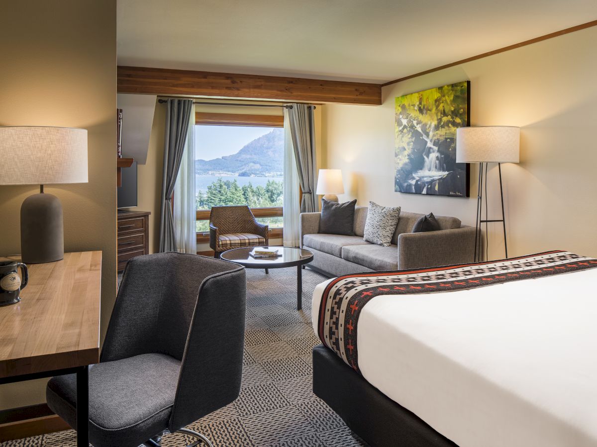 Hotel room with a bed, seating area by a window, lamps, and a desk; cozy decor and mountain view outside, bright and inviting.