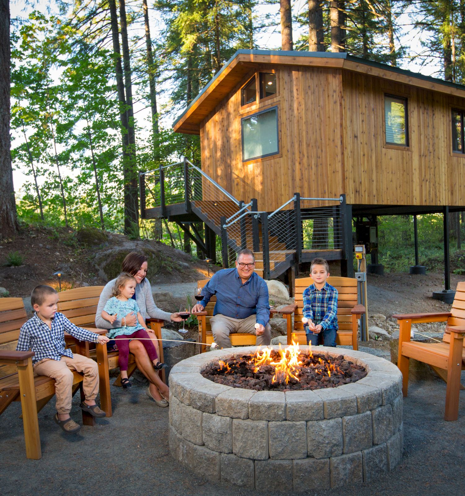 A family sits around a fire pit with wooden chairs in a forest clearing, a small wooden cabin on stilts in the background.