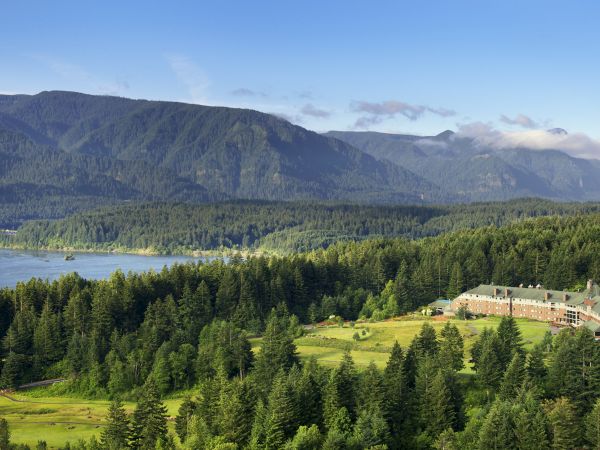 A riverside forest retreat sits below misty mountains, with a steel bridge crossing the water under a clear blue sky.
