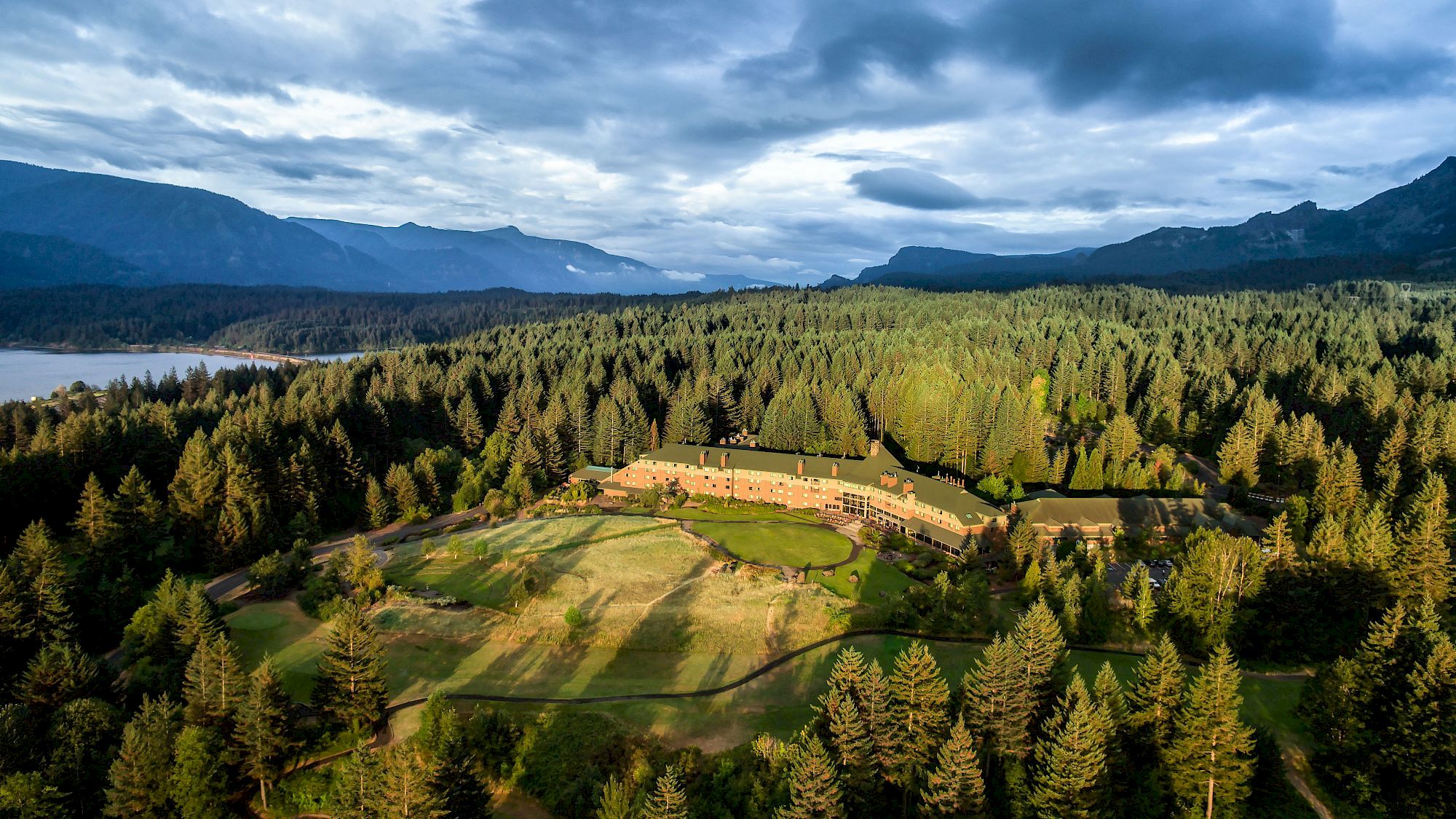 A scenic aerial view of a forested landscape with a long building complex, rolling hills, and a distant lake under a dramatic cloudy sky.