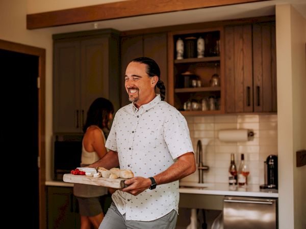 A smiling man carries a tray of food into a kitchen, while a woman in the background chats near the counter, warm home setting.