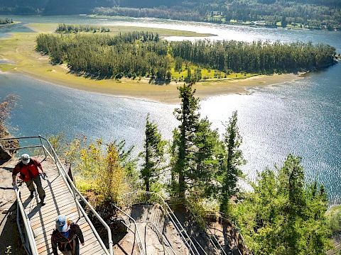 People hike on a wooden boardwalk along a river lake, with forested island and towering mountains in the background.