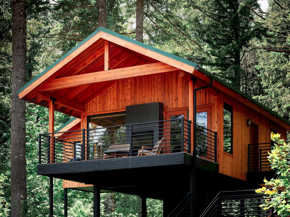 A wooden elevated cabin in a forest, with a balcony and stairs, surrounded by tall trees, under a sunny sky.