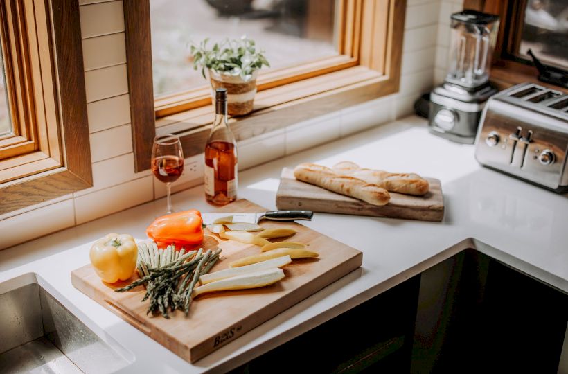 Cheese, charcuterie, and bread on a wooden board by the window, with wine, herbs, and a lit candle; kitchen counter and toaster in view.