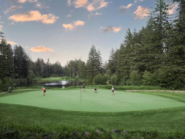 A group of people are playing golf on a green surrounded by trees and a lake under a sky with clouds.