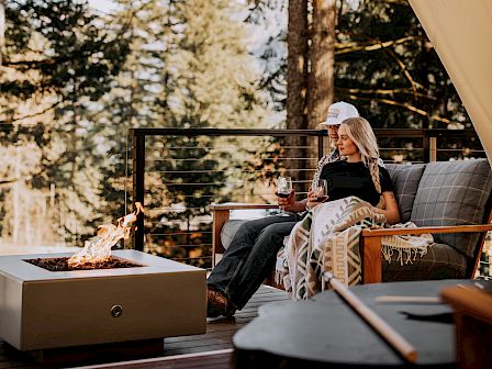 A woman relaxes on an outdoor patio under a large canvas tent, sipping wine by a crackling fire.