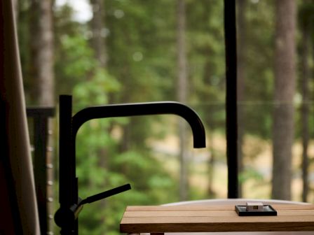 A wooden worktable with a black metal arm sits by a window, overlooking green trees in the blurred background.