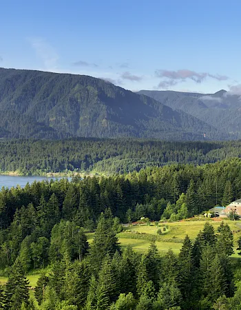 A scenic mountain valley with a river, dense forest, a long bridge, and a large resort building under blue skies.