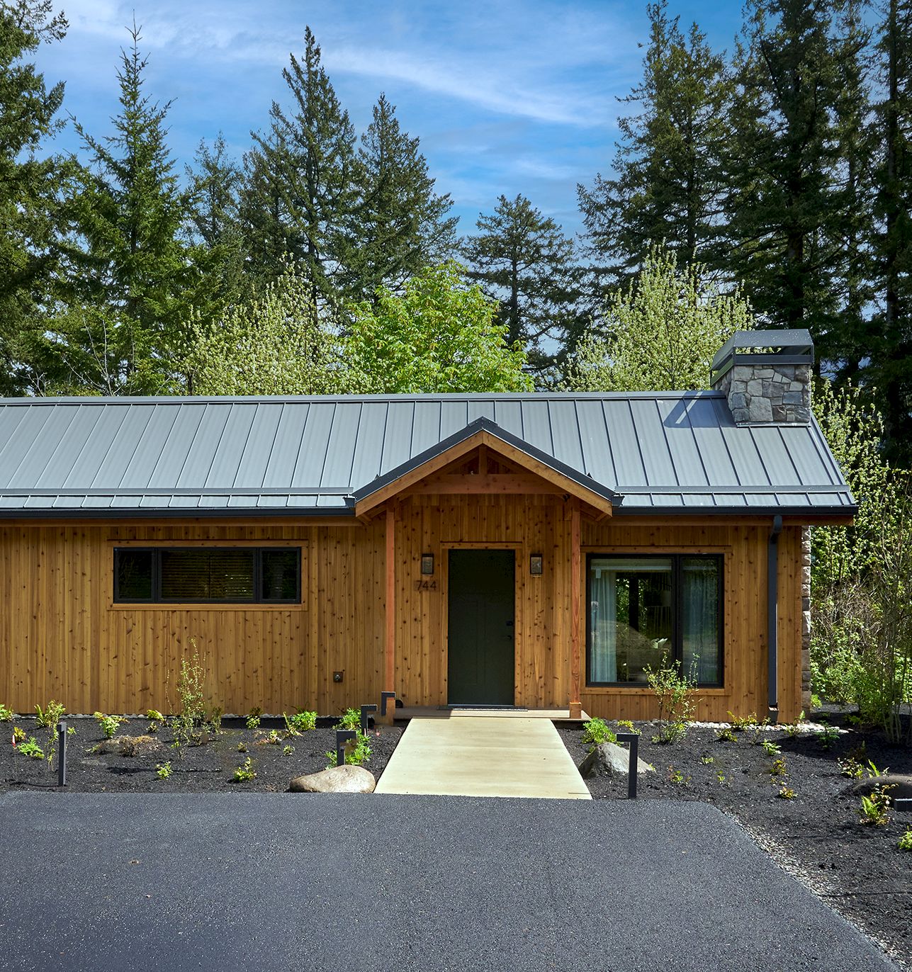 A modern wooden house with a silver metal roof and stone chimney sits among trees, with a front path and driveway leading to the door.