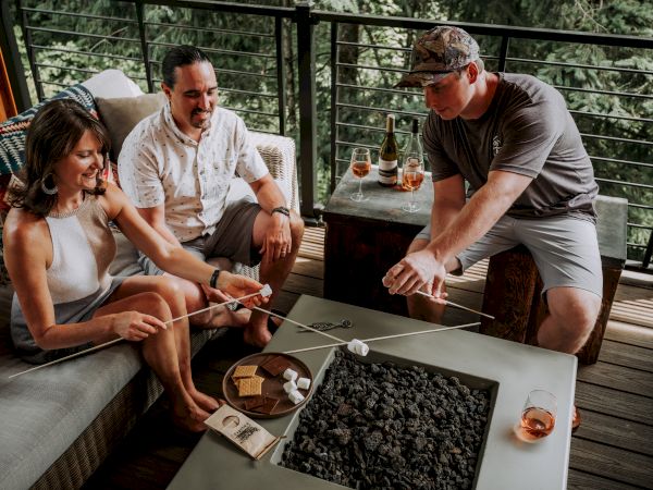 Friends roasting marshmallows and snacking on chocolate on a balcony, with wine glasses and bottles on the table, enjoying outdoors.