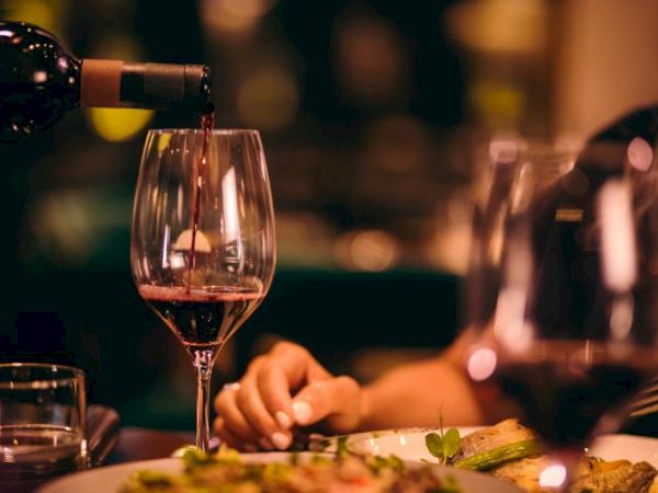 A person pours red wine into a glass, with a dinner plate and another glass visible on the table in a dimly lit setting.