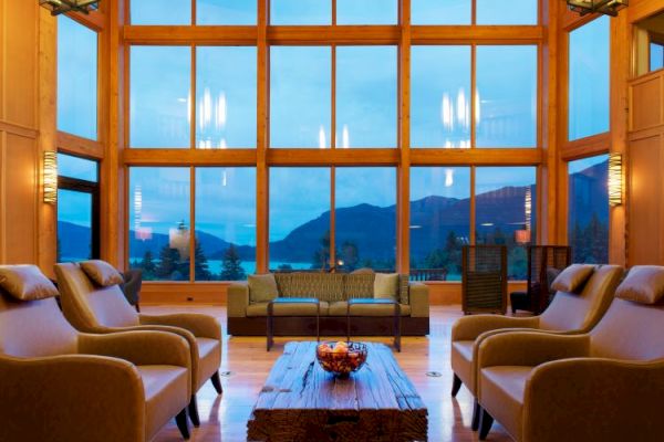 A cozy living room with large windows, offering a mountain and lake view, featuring armchairs and a wooden coffee table.