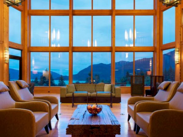 A cozy living room with large windows, offering a mountain and lake view, featuring armchairs and a wooden coffee table.