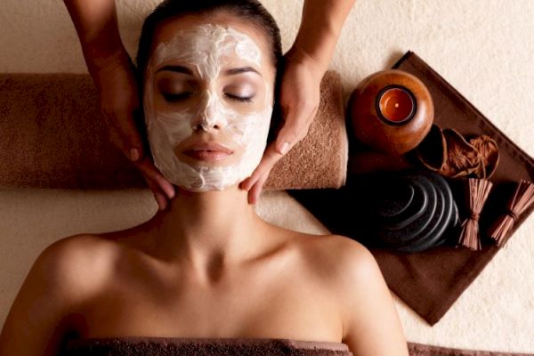 A person is receiving a facial treatment with a cream mask applied, surrounded by spa items like stones and a lit candle.