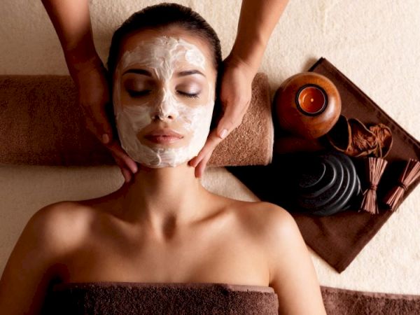 A person is receiving a facial treatment with a cream mask applied, surrounded by spa items like stones and a lit candle.