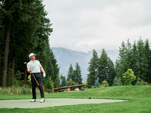 A golfer stands on a tee in a lush, forested course, mountains in the distance, ready to swing as the sun peeks through clouds.