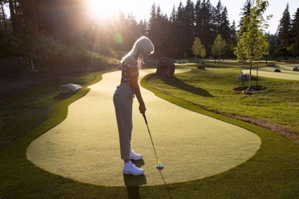 A person is playing golf on a putting green surrounded by trees and rocks, illuminated by the setting sun in a tranquil setting.
