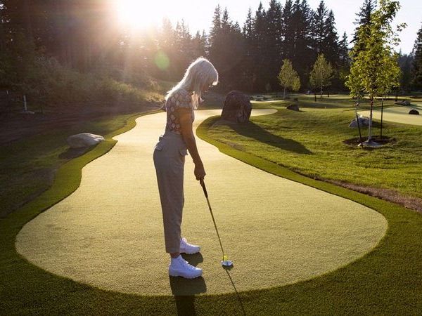A person is playing golf on a putting green surrounded by trees and rocks, illuminated by the setting sun in a tranquil setting.