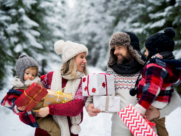 A family, dressed warmly in winter clothes, stands in a snowy forest holding wrapped gifts, smiling and looking at each other.