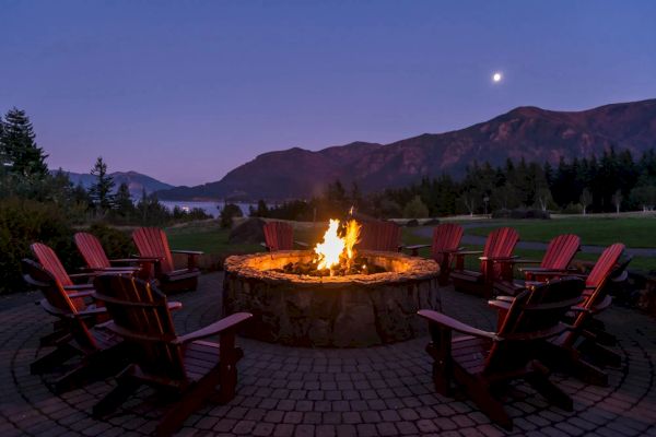 A cozy outdoor fire pit with Adirondack chairs, set against a twilight mountain backdrop and crescent moon in the sky.