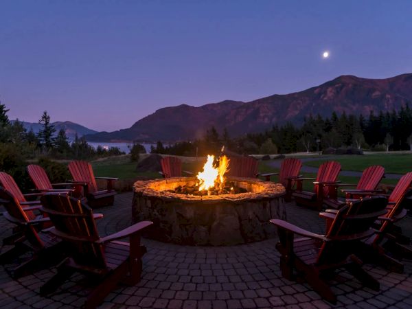 A cozy outdoor fire pit with Adirondack chairs, set against a twilight mountain backdrop and crescent moon in the sky.