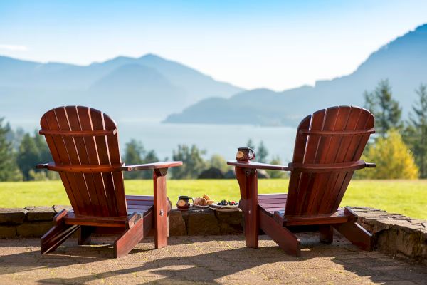 Two wooden chairs overlook a scenic view of mountains and a lake, with trees and clear skies, creating a peaceful outdoor setting.