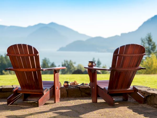 Two wooden chairs overlook a scenic view of mountains and a lake, with trees and clear skies, creating a peaceful outdoor setting.