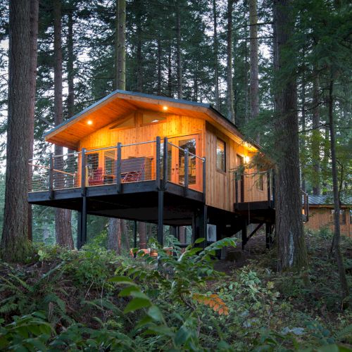 A glowing wooden treehouse cabin with a wraparound deck sits high among tall forest trees, lit at dusk.