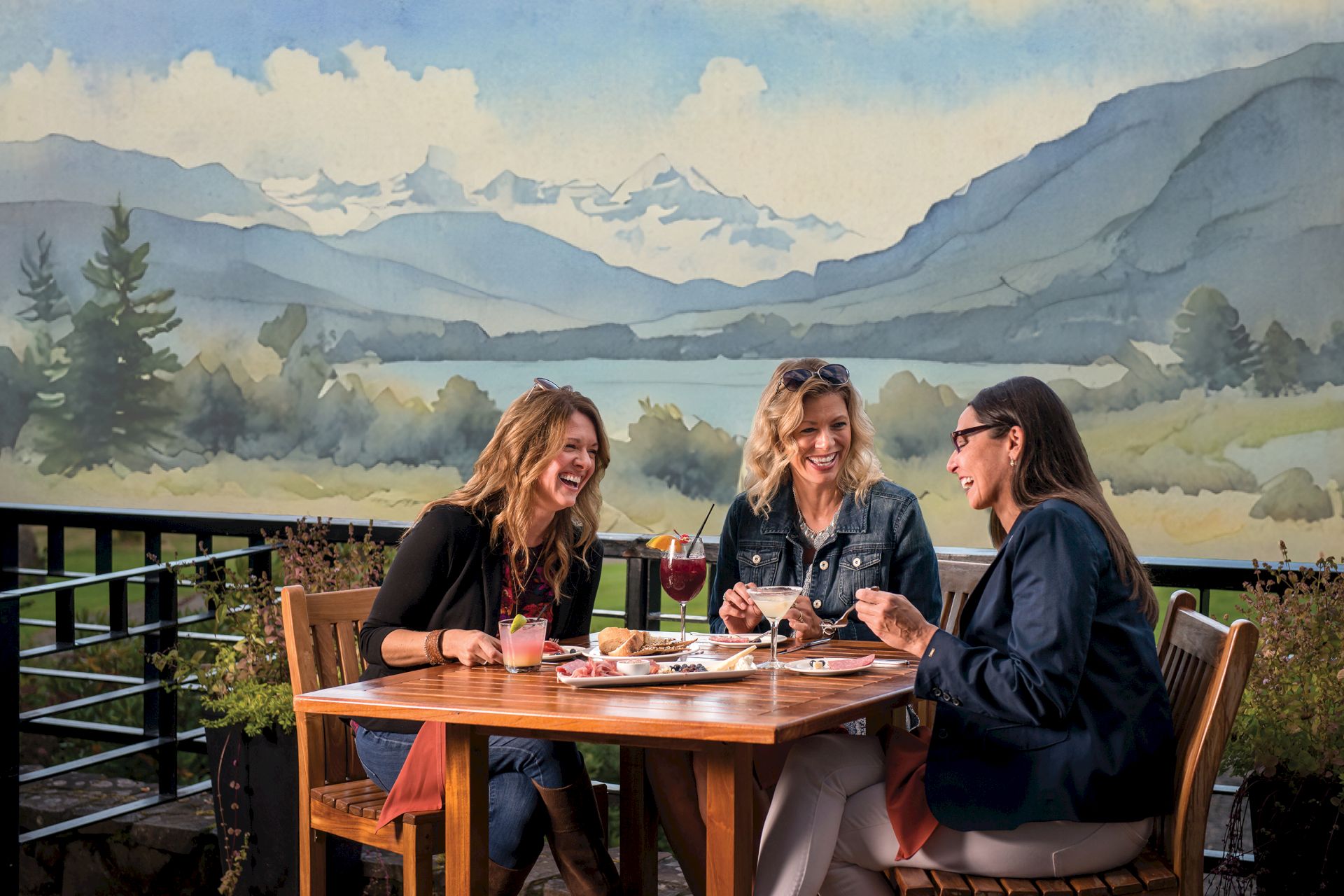 Three people enjoy drinks and appetizers at a wooden outdoor table, with a scenic mountain mural in the background.