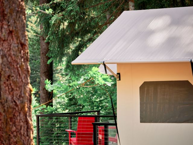 A white cabin with a canvas awning sits on a deck in a forest, with red chairs visible through the railing.