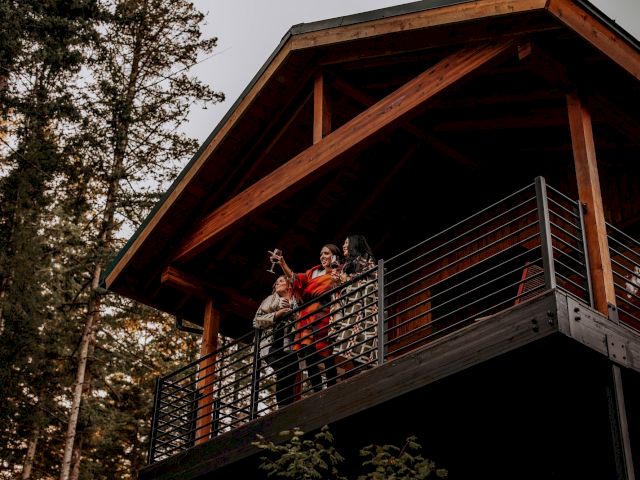 People toast on a wooden cabin balcony, holding drinks in front of a slanted timber roof among trees.