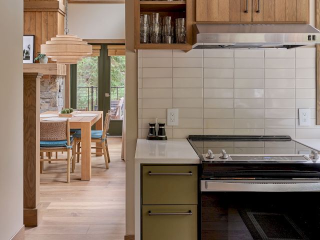 Warm modern kitchen with wood cabinetry, open shelves of glass jars, tile backsplash, and a stainless gas stove in view.