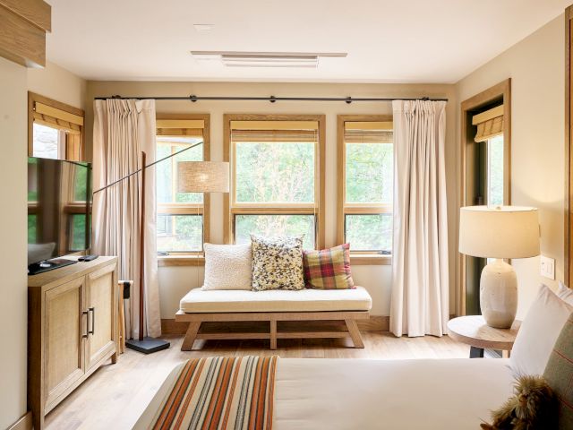 A bright bedroom with wooden windows, white curtains, a cushioned bench by the glass, and patterned pillows and rugs.