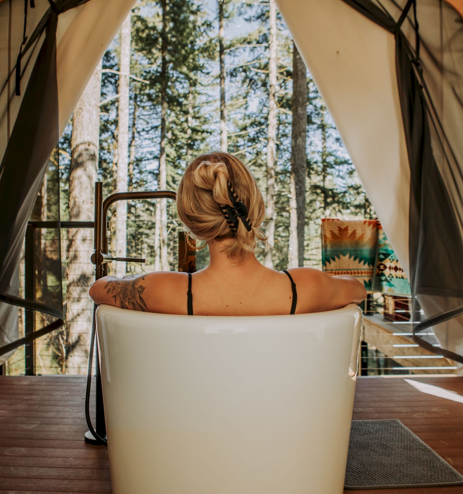 A woman relaxes in a white hot tub inside a cozy cabin, looking out at a forest through open curtains.