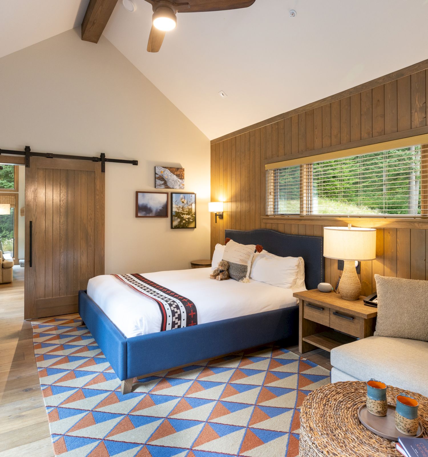 A cozy modern bedroom with a blue bed, wood-paneled walls, large window blinds, lamps, and a patterned rug.