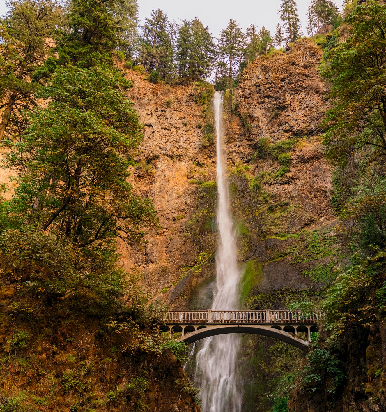 A tall waterfall plunges down a rocky forest canyon beside an arched stone bridge, with mist and trees framing the scene.