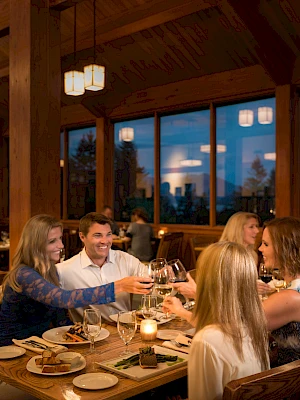 A group of people enjoying a meal at a cozy restaurant, clinking glasses at a wooden table with dim lighting and large windows.