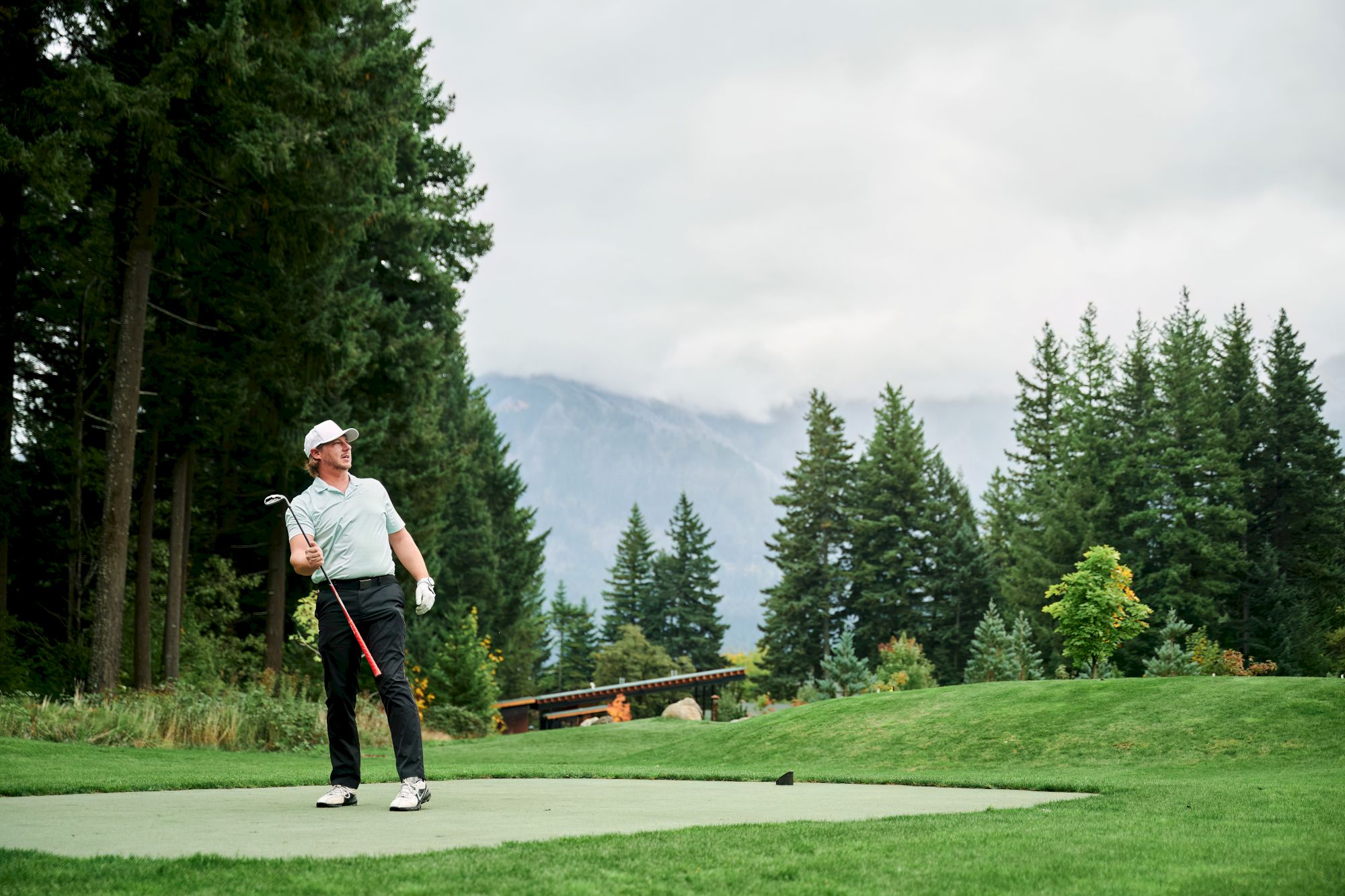 A golfer in a cap swings a club on a green course surrounded by pine trees and distant mountains under cloudy skies.