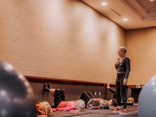 An indoor training session shows a woman standing and gesturing as people on the floor and chairs watch her.