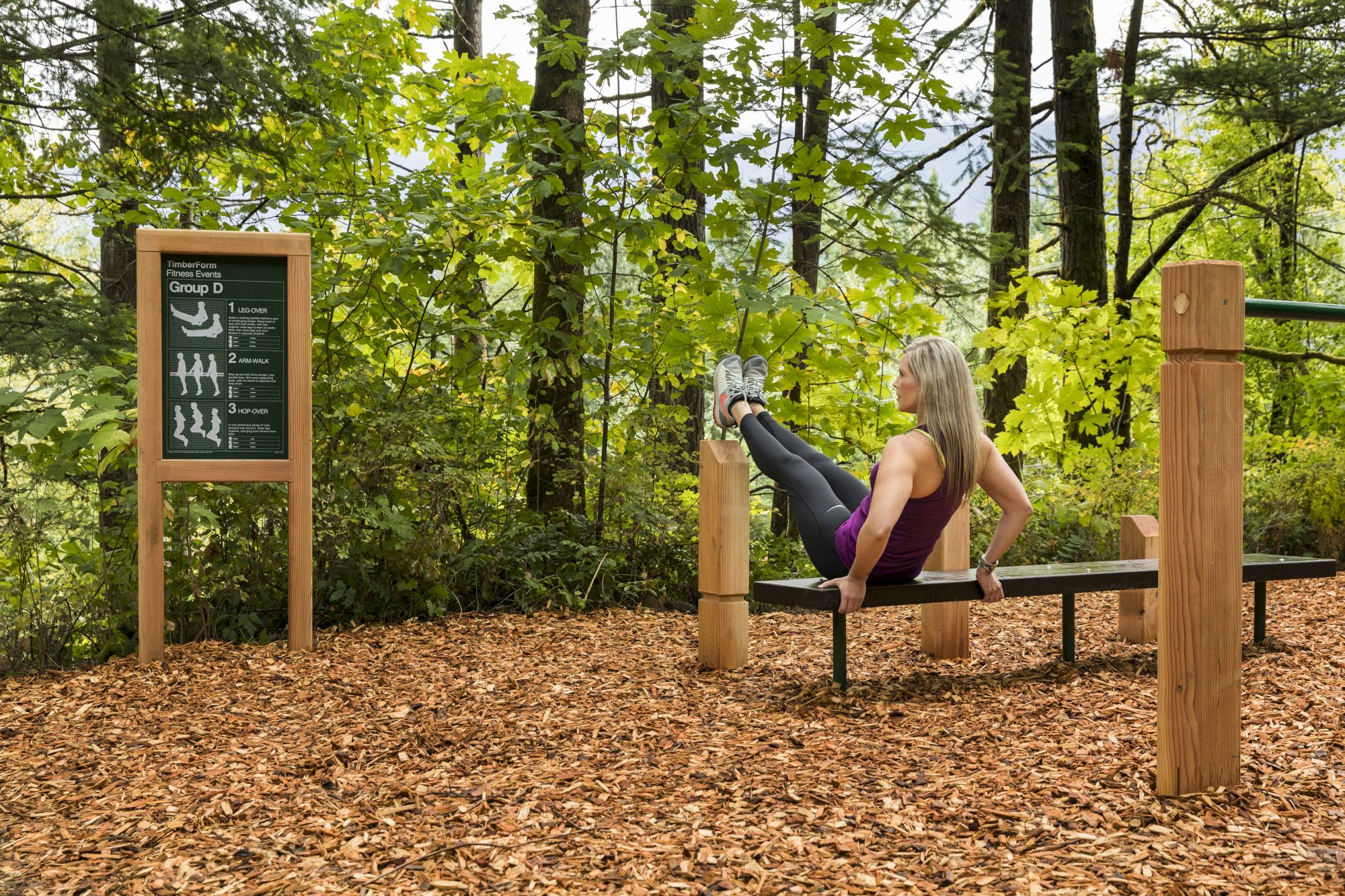In a forest workout area, a woman does exercises on a wooden bench while a sign shows guided moves.