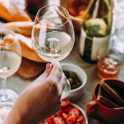A hand clinks a wine glass while a table of food and plates sits around it, like a celebratory dinner with champagne.