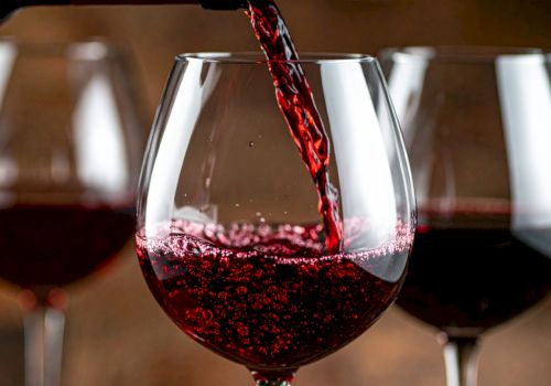 A stream of red wine pours into a glass, with other wine glasses in the background.