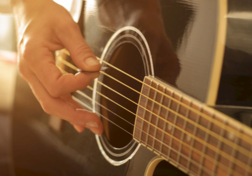 A person&rsquo;s hand plucks the strings of an acoustic guitar in a warm, close-up shot.