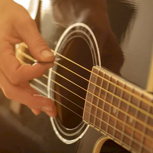 A person’s hand plucks the strings of an acoustic guitar in a warm, close-up shot.