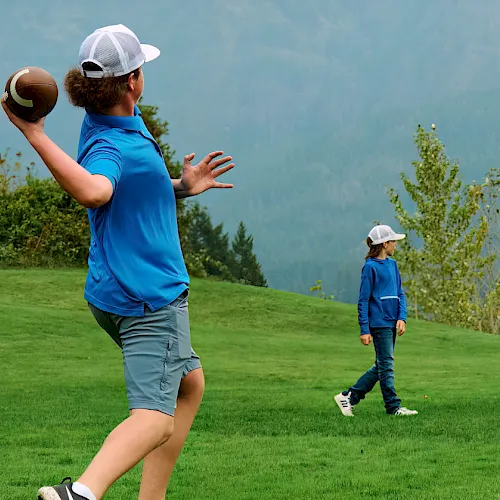 A woman and child in blue play football on a grassy field with mountains in the background.
