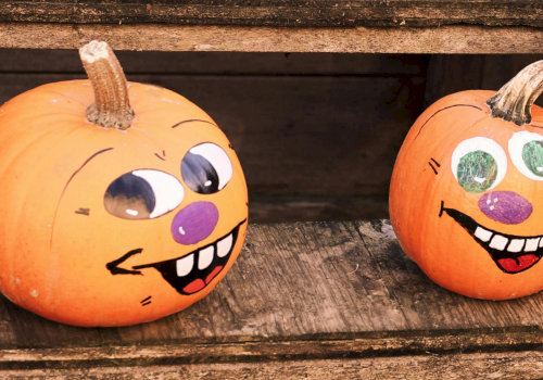 Two Halloween pumpkins carved with silly faces sit on a wooden surface, one with a stem and the other smiling wide.