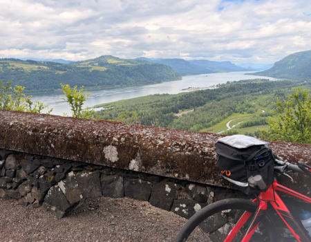 A red motorcycle rests beside a stone lookout wall overlooking a winding river and lush green hills under cloudy skies.