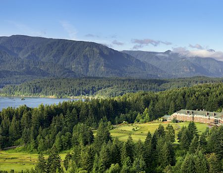 A scenic valley with dense evergreen forests, a river, a metal bridge, and a lodge beneath towering mountains and drifting clouds.