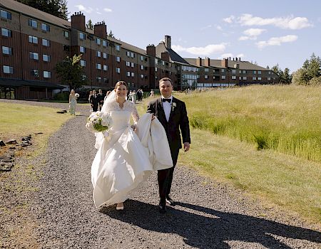 A bride and groom in wedding attire walk along a gravel path by a lodge, surrounded by guests and greenery.