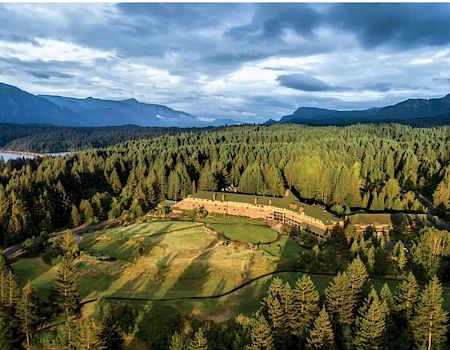 A scenic mountain valley with a large hotel/resort building surrounded by dense pine forests under cloudy skies.